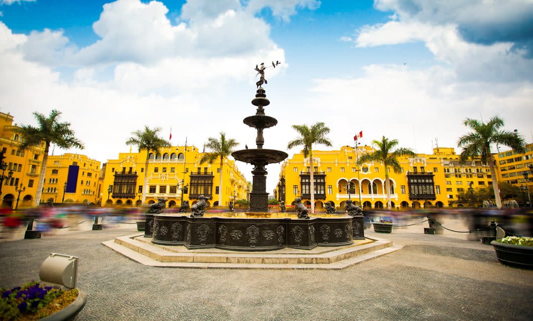 A large, black ornate fountain stands in the center of a plaza surrounded by tall palm trees and bright yellow colonial buildings under a partly cloudy blue sky—an iconic scene on any Deluxe Tour of South America. People are visible around the square.
