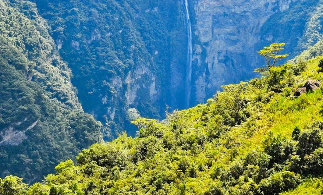 Lush green forest covers sloping hills in the foreground, with steep cliffs and the narrow, tall Gocta Waterfalls cascading down the rocky background in a vibrant Chachapoyas mountain landscape.