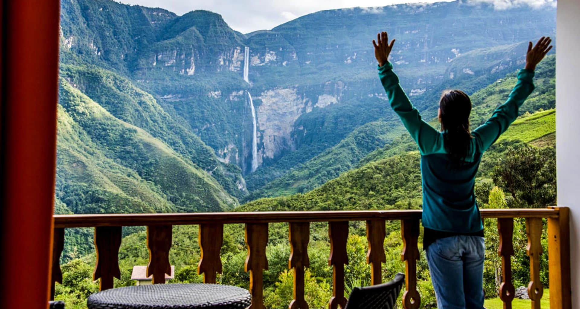 A person stands on a balcony with arms raised, overlooking the lush green mountains near Gocta Waterfalls, close to Chachapoyas. The vibrant scene suggests awe and a deep connection with nature’s beauty.