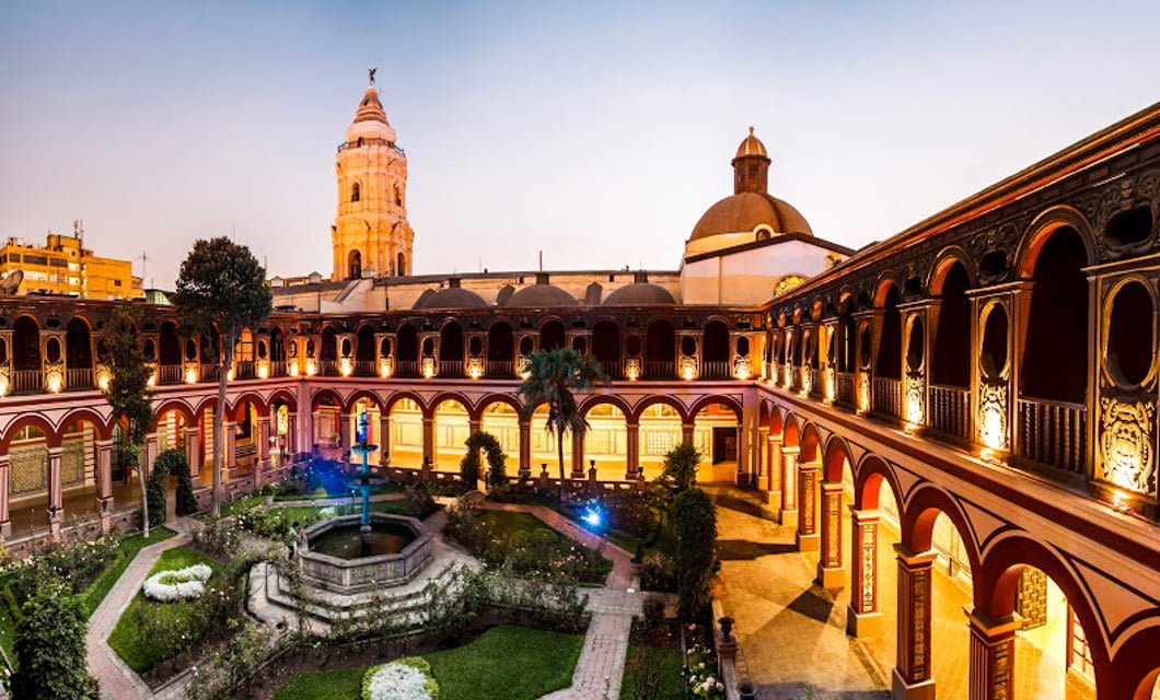 A colonial-style courtyard with red arches, a central fountain, and gardens, surrounded by a two-story cloister—perfect for a relaxing tour. In the background, church domes and a tall bell tower glow at sunset.