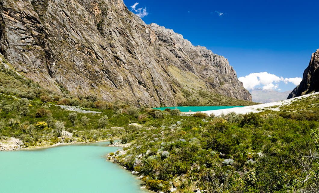 Turquoise river winding through lush green vegetation, flanked by steep rocky mountains of the Cordillera Blanca under a bright blue sky with scattered clouds—an iconic scene from a classic Peru Trek near Huaraz.