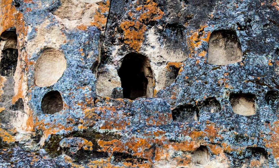 A rocky cliff face with numerous carved openings and niches, covered in patches of orange, blue, and black lichen; its weathered surface hints at secrets waiting to be discovered on a Cajamarca Archaeology Tour.