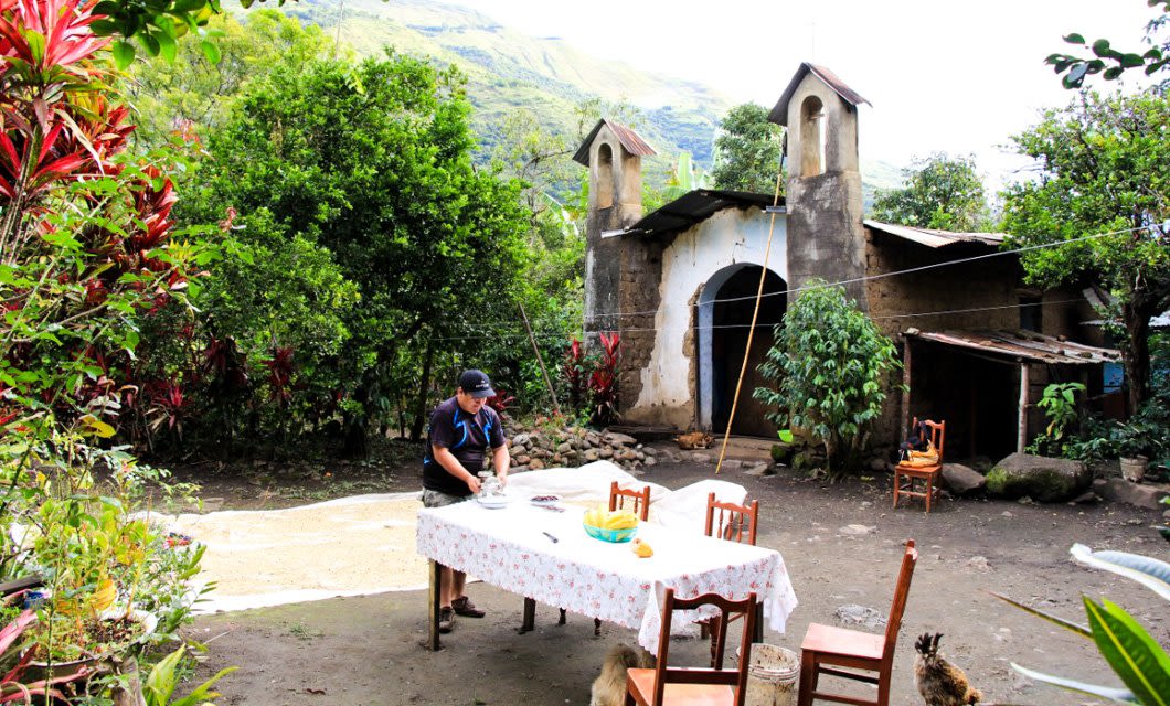 A person sets a table with a floral tablecloth outdoors, surrounded by chairs, lush greenery, and red plants. An old, rustic chapel with twin bell towers stands in the background—a perfect scene for a chocolate tour in Peru under a bright sky.