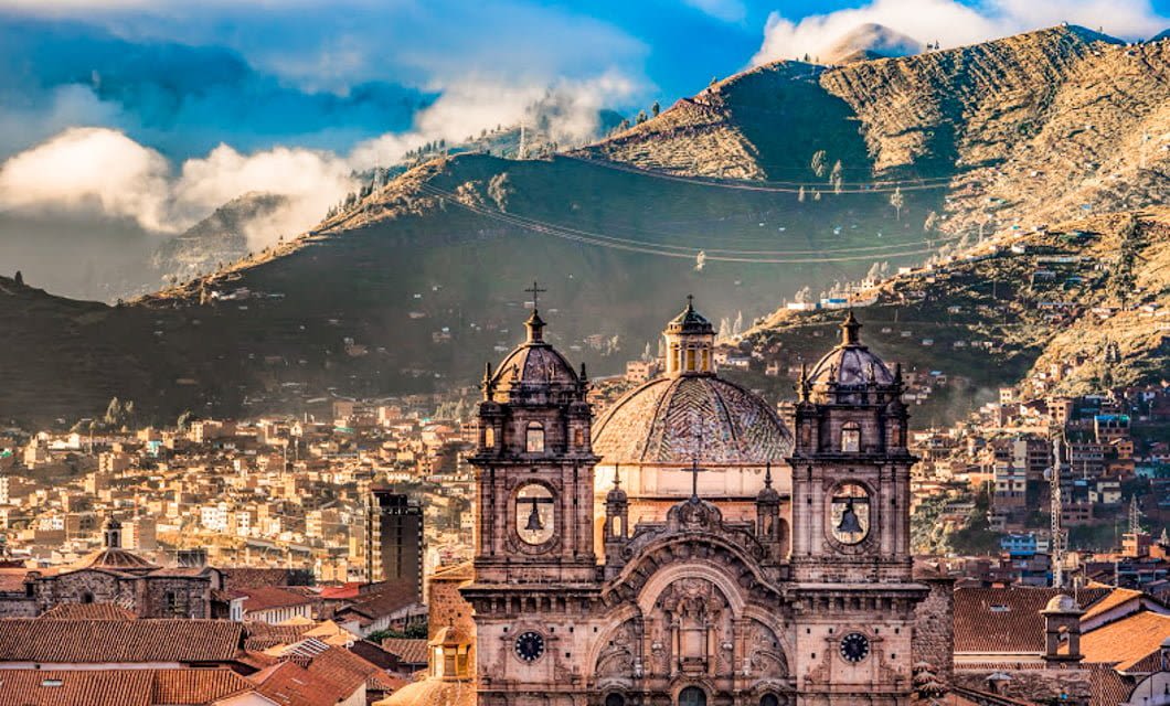 A historic cathedral with twin bell towers stands in the foreground, reflecting Peru's rich heritage, backed by a cityscape and green mountains under a blue sky with clouds.