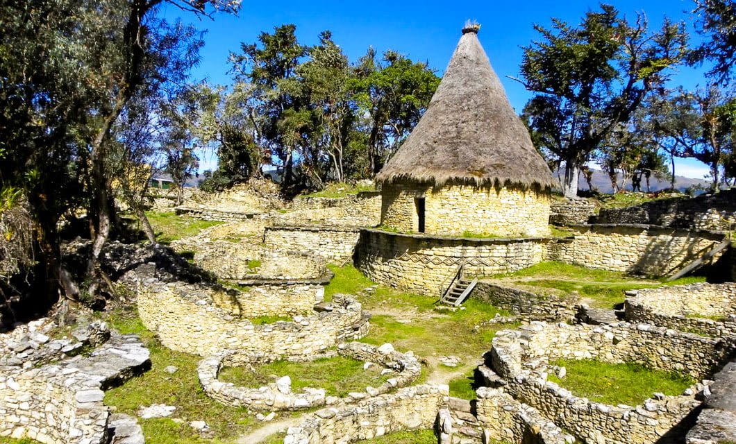 Ancient stone ruins with circular walls and a large round building with a conical thatched roof, reminiscent of Kuelap, surrounded by trees and greenery under a clear blue sky.