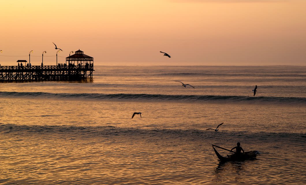 A silhouette of a person in a small boat on the ocean at sunset, with seagulls flying and a wooden pier with a gazebo extending over the water in the background, evokes the serene coastlines near Trujillo and ancient wonders like Chan Chan.