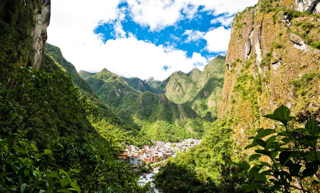 A small town nestled in a lush green valley along the Salkantay Trek, surrounded by steep, forested mountains under a bright blue sky with scattered clouds.