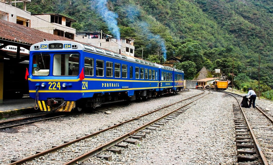 A blue and yellow train labeled 224 sits on tracks beside a mountainous landscape in Peru, with smoke coming from its engine. Nearby, a person stands next to the rails as buildings and greenery highlight this scenic train tour.