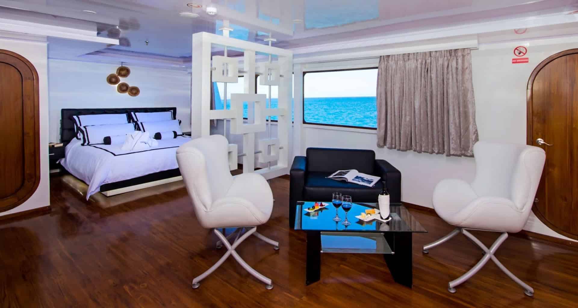 Modern suite aboard the M/C Petrel Galapagos Cruise with wood floors, a bed with white linens, a seating area with two white chairs, a black chair, a glass table with drinks and snacks, and a large window showcasing ocean views.