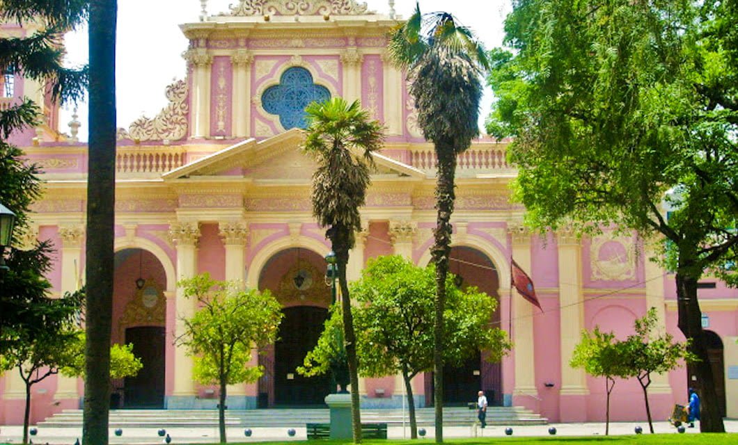 Historic pink and yellow cathedral in Salta, Argentina, with arched entrances, ornate details, and a large central rose window, surrounded by palm trees and greenery—a must-see on any adventure tour, with people walking in front.