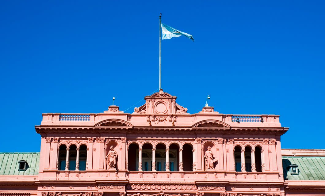The top of a pink historic building with arched windows and statues, flying the Argentine flag against a clear blue sky—a stunning sight to admire during your South America Golf Tour.