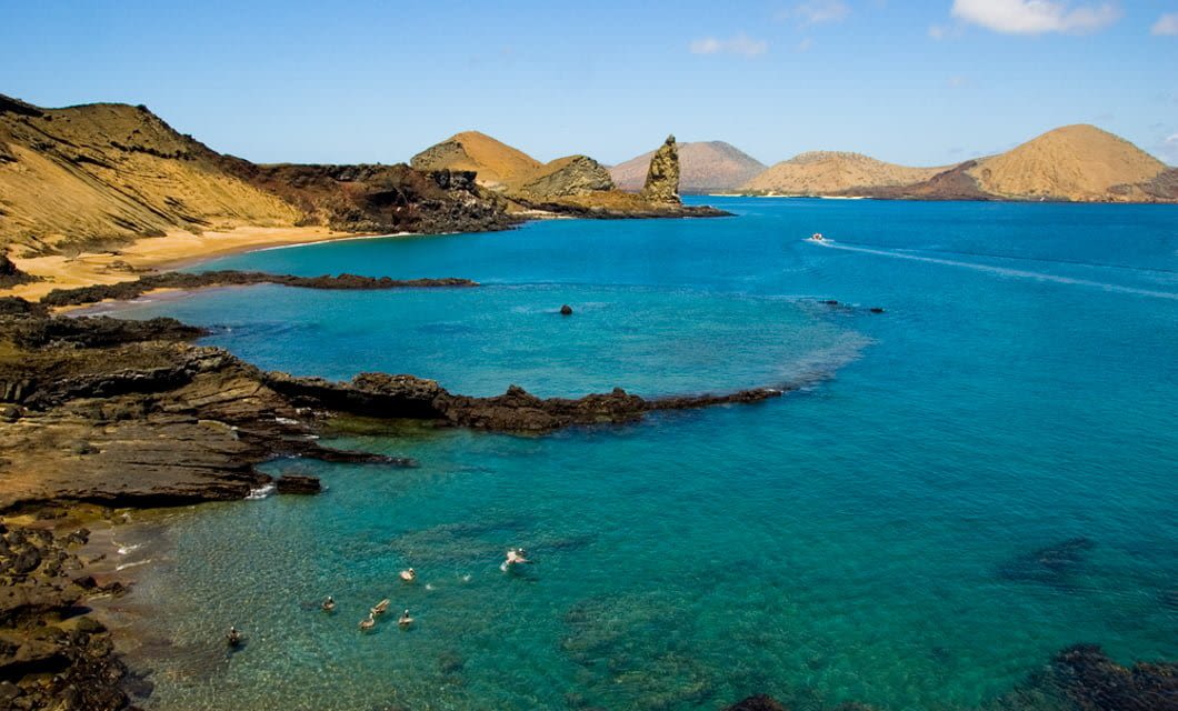 A clear blue bay with rocky shores and distant hills under a bright sky. Several swimmers enjoy the shallow water near the shoreline, where the M/C Petrel offers a unique Galapagos cruise experience amid sandy beaches and rugged terrain.