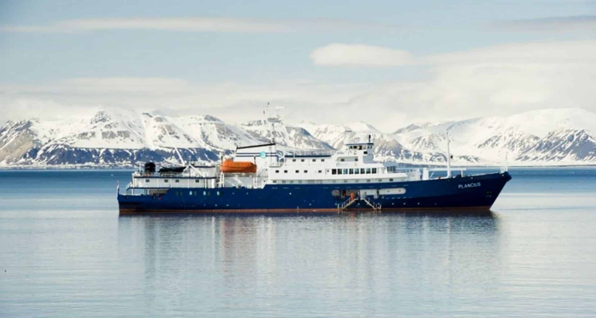 A blue and white ship, the Plancius, is anchored on calm water with snowy mountains in the background under a partly cloudy sky, capturing the serene beauty of an Antarctica Cruise departing from Ushuaia.