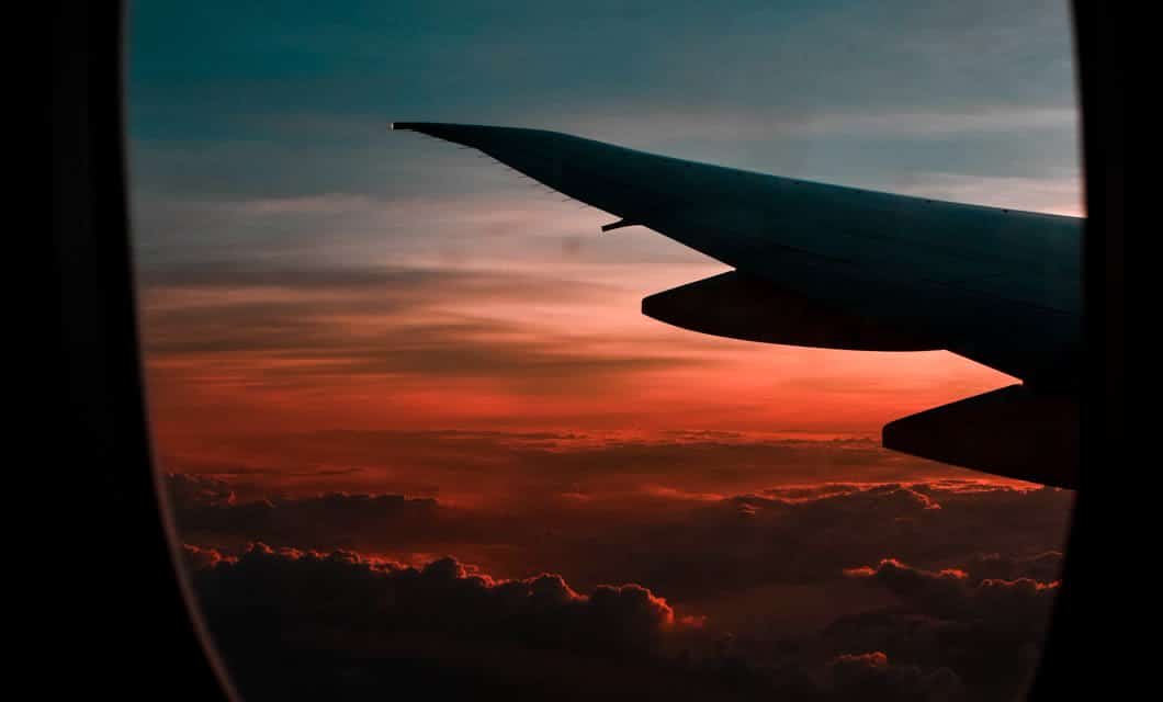 View from an airplane window showing a wing silhouetted against a dramatic sunset sky, clouds below glowing in shades of orange, pink, and blue—a perfect vista on your Guided Group Tour to Patagonia.