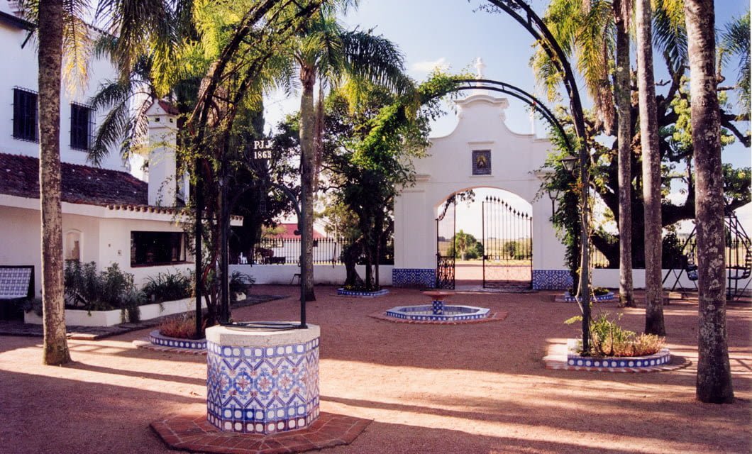 Courtyard with Spanish colonial architecture, featuring tiled fountains and a well, palm trees, archways, and a white gated entrance in the background—evoking the timeless charm of Montevideo and classic Estancias of Uruguay under a clear sky.