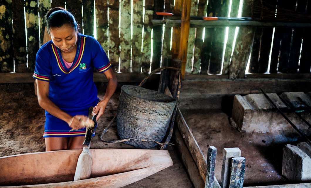 A woman in a blue dress processes food with a wooden tool inside a rustic building, as sunlight filters through the walls—a glimpse into authentic life often experienced on Anakonda Amazon Cruises, where luxury river cruise adventures meet tradition.