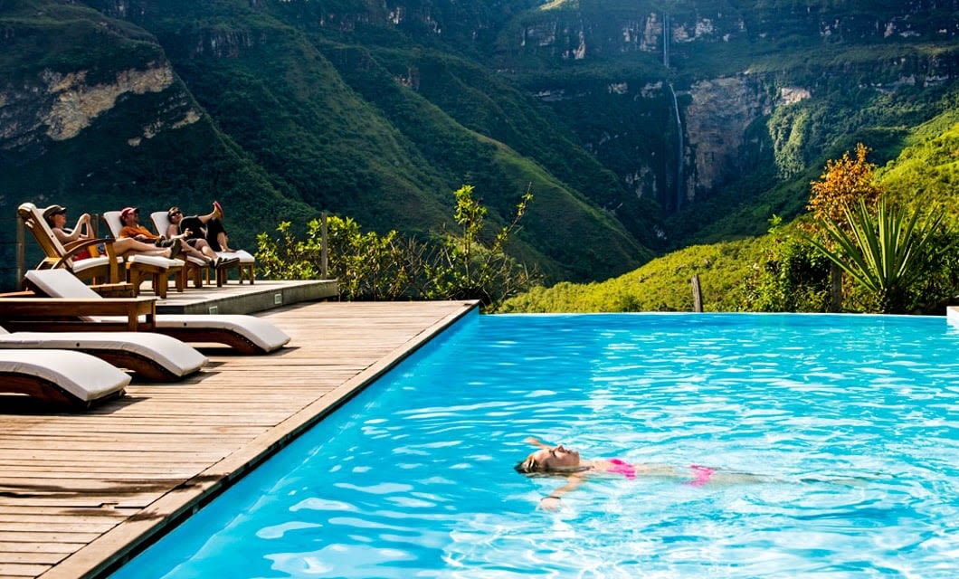 A woman floats in a bright blue pool, surrounded by lush green mountains near Chachapoyas. Several people lounge poolside under the sun, enjoying the peaceful and scenic atmosphere inspired by Gocta Waterfalls.