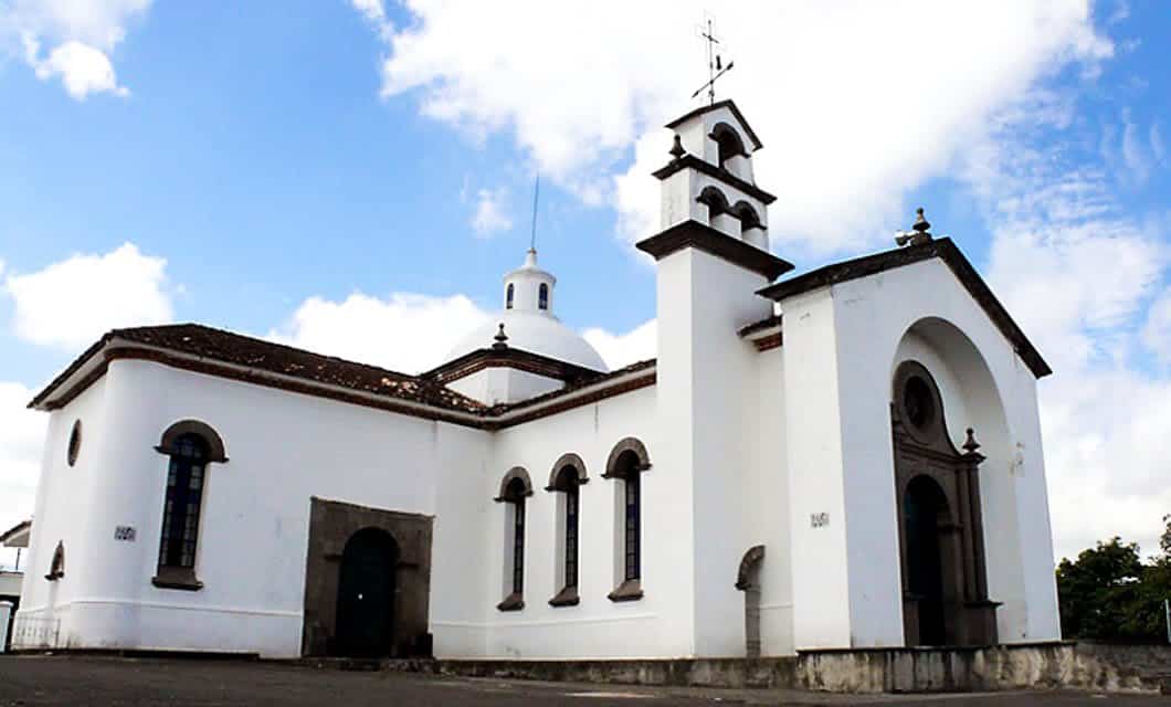White colonial-style church with arched windows, a bell tower, and a dome set against a blue sky. Dark trim and a cross crown the tower. A highlight on any Colombia Archaeology Tour near the famed San Agustín Ruins.