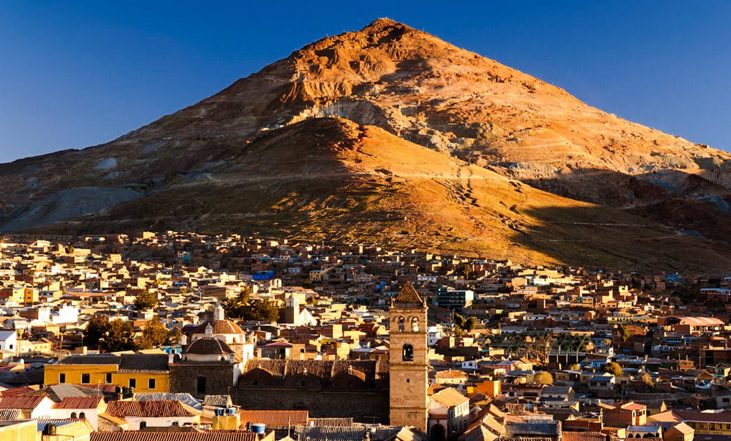 A golden mountain looms over the city of Potosí, Bolivia, with clustered colorful buildings below, bathed in warm sunlight and clear blue sky above—a must-see stop on any La Paz tour or journey to Uyuni.