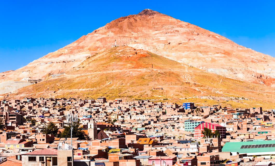 A city with densely packed buildings sits at the base of a large, reddish-brown mountain under a clear blue sky, reminiscent of Potosi near the edge of the Atacama Desert.