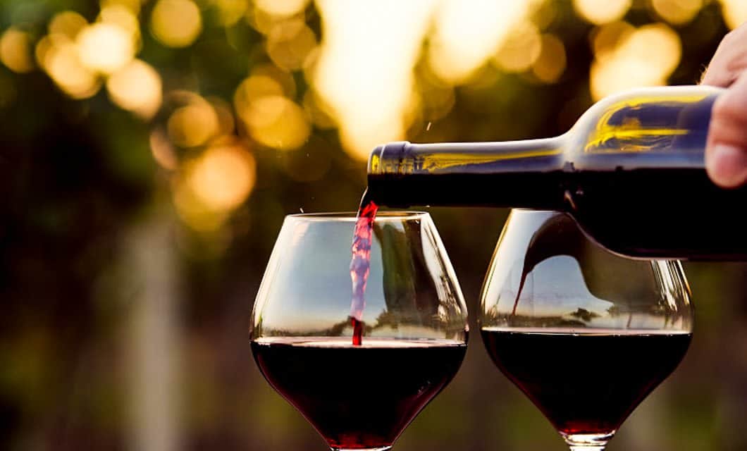A hand pours red wine from a bottle into one of two glasses, with a blurred outdoor background and warm sunset lighting—perfect for a Mendoza honeymoon.
