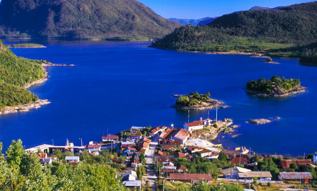 A small town with red-roofed houses sits on the edge of a deep blue lake in Patagonia, surrounded by green hills and mountains under a clear sky. Small islands and patches of greenery dot the lake, reminiscent of the scenic Chilean Fjords.