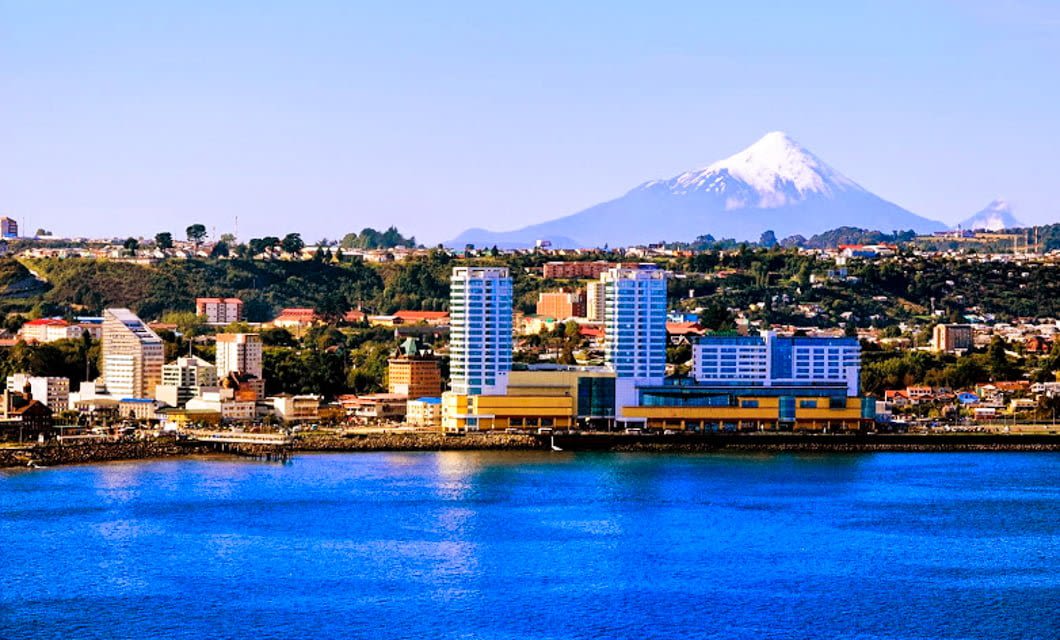 A coastal city with modern buildings by the waterfront, colorful houses on hills, and a snow-capped volcano in the background under a clear blue sky—reminiscent of Puerto Varas in Chile.