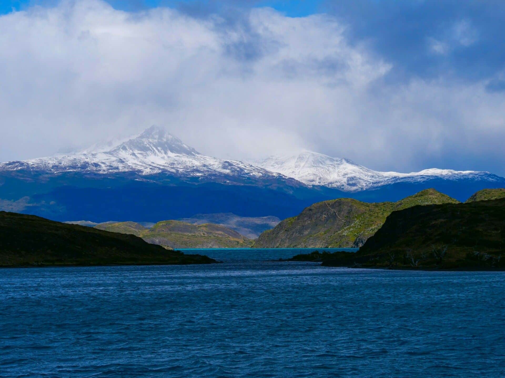 A tranquil blue lake with dark green hills in the foreground and a range of snow-capped mountains beneath a cloudy sky in the background—discover these Patagonia essentials on a guided group tour.