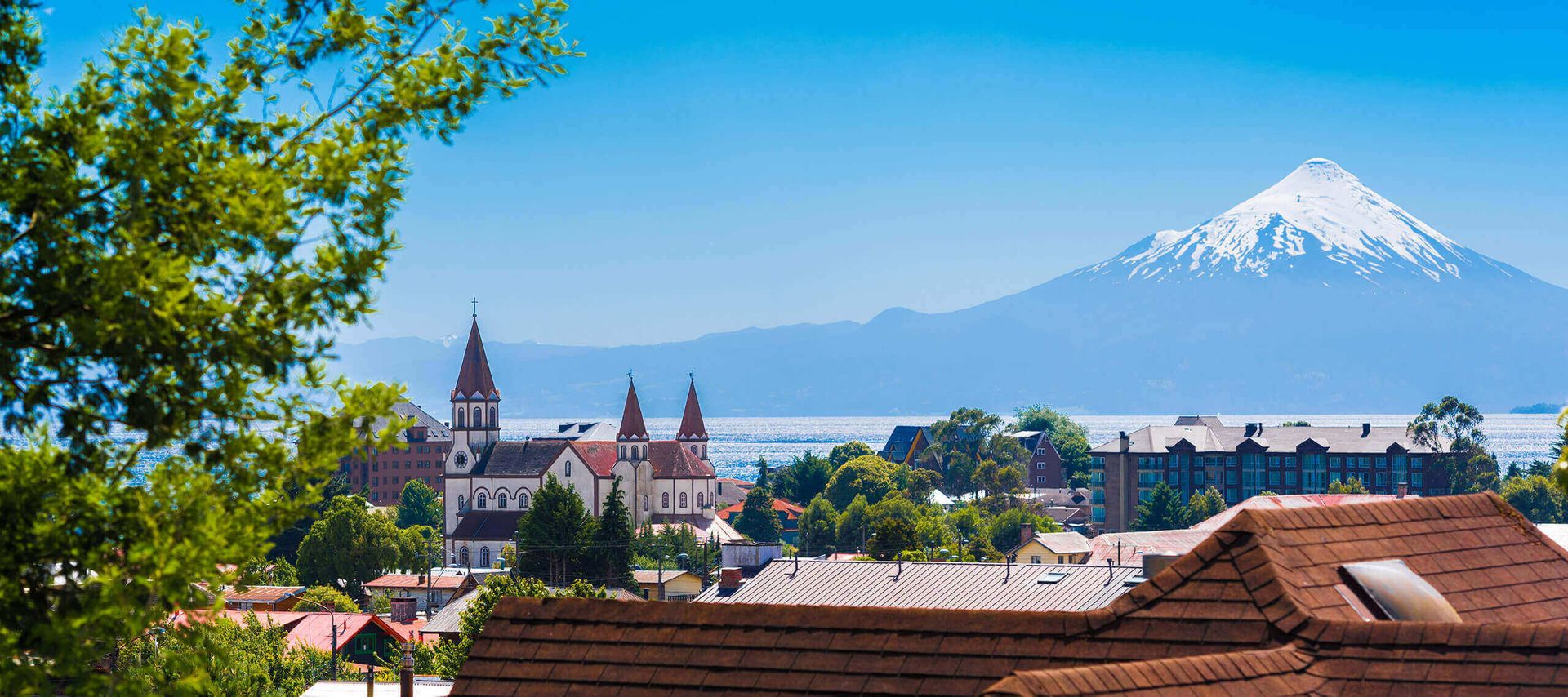 A scenic view of a town with red-roofed buildings and a church, set against a backdrop of a snow-capped volcano and blue sky, with green foliage framing the left side—an iconic scene from the Chile Lake District.