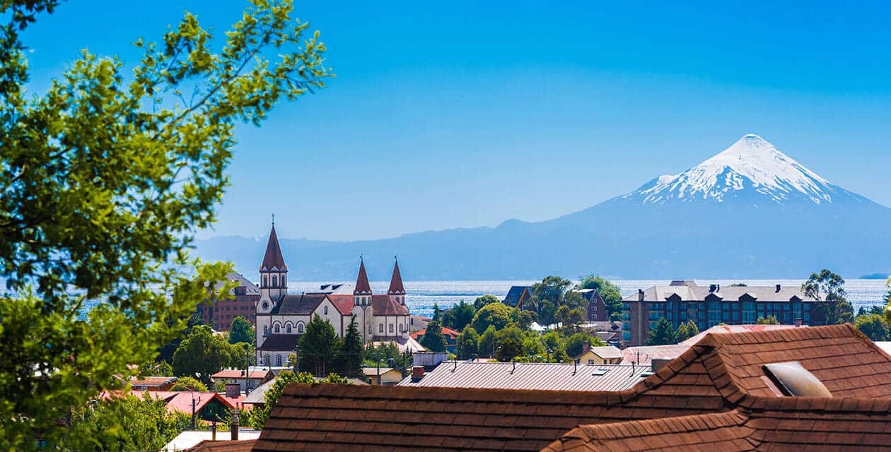 A scenic view of a town with red-roofed buildings, a church with pointed towers, lush greenery, and a snow-capped volcano in the background, all set near serene lakes under a clear blue sky.
