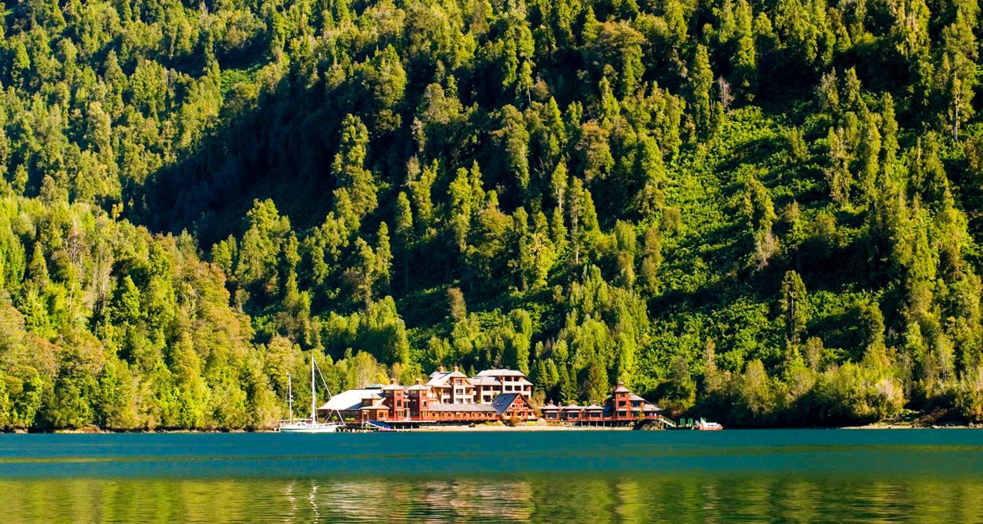 A large wooden eco resort, Puyuhuapi Lodge, sits on the edge of a calm lake in Chile, surrounded by dense green forest and steep hills under bright sunlight. A sailboat is docked nearby.