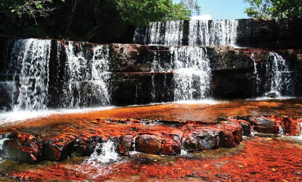 A multi-tiered waterfall flows over dark rocks into a shallow pool, its vibrant orange-red hues contrasting with the white water—an iconic sight along the Mount Roraima Trek, surrounded by lush green foliage.