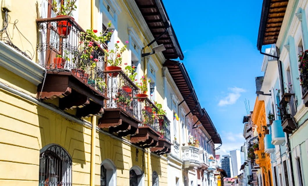 Colorful colonial-style buildings with wrought-iron balconies decorated with potted flowers line a narrow street in Colonial Quito, beneath a bright blue sky, evoking the charm of this vibrant, historic urban area.