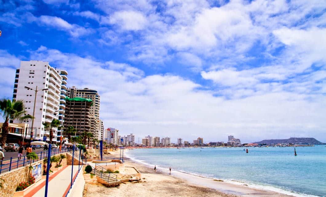 A coastal cityscape with tall modern buildings lining a sandy beach under a bright blue sky with scattered clouds. Gentle waves touch the shore, and a few people stroll along the beach—an inviting scene on an Ecuador Tour.