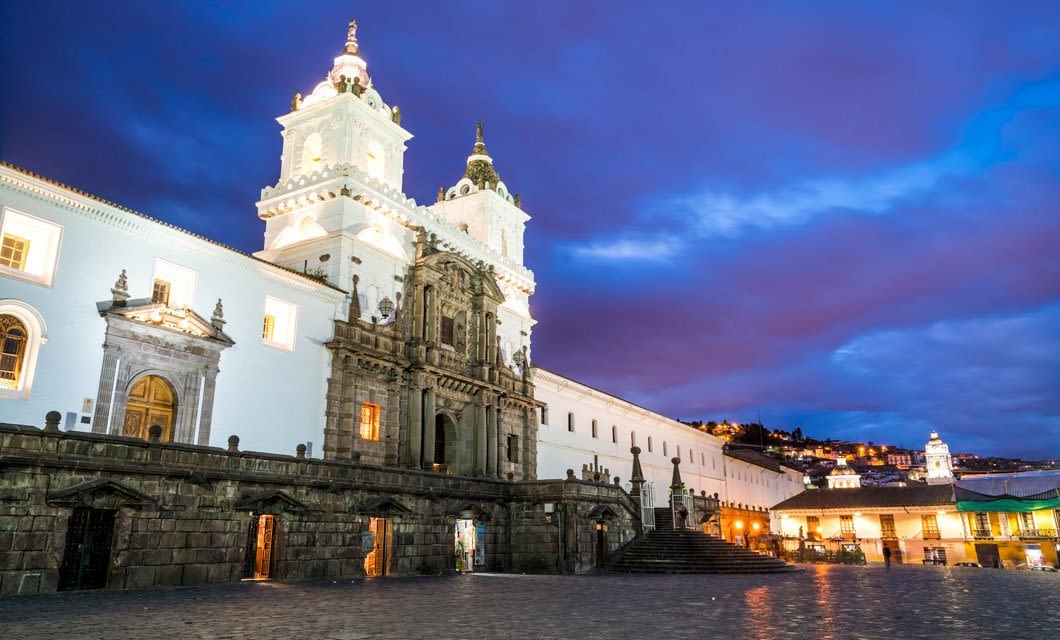 Historic white stone church with ornate twin bell towers and a cobblestone plaza, glowing under evening lights and a dramatic sky in Quito, the perfect start to any memorable Ecuador tour.