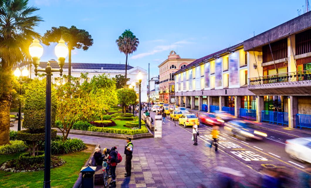City street at dusk in the Heart of the Andes, with blurred car lights, people walking on sidewalks, a green park with trees and lampposts on the left, and colorful buildings on the right under a blue sky.