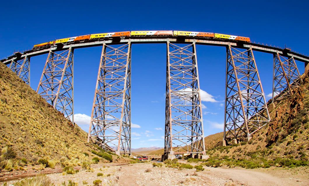 A brightly colored train travels across a tall steel viaduct bridge in Salta, Argentina, spanning a dry valley with hills and sparse vegetation under a clear blue sky—perfect for an unforgettable adventure tour.