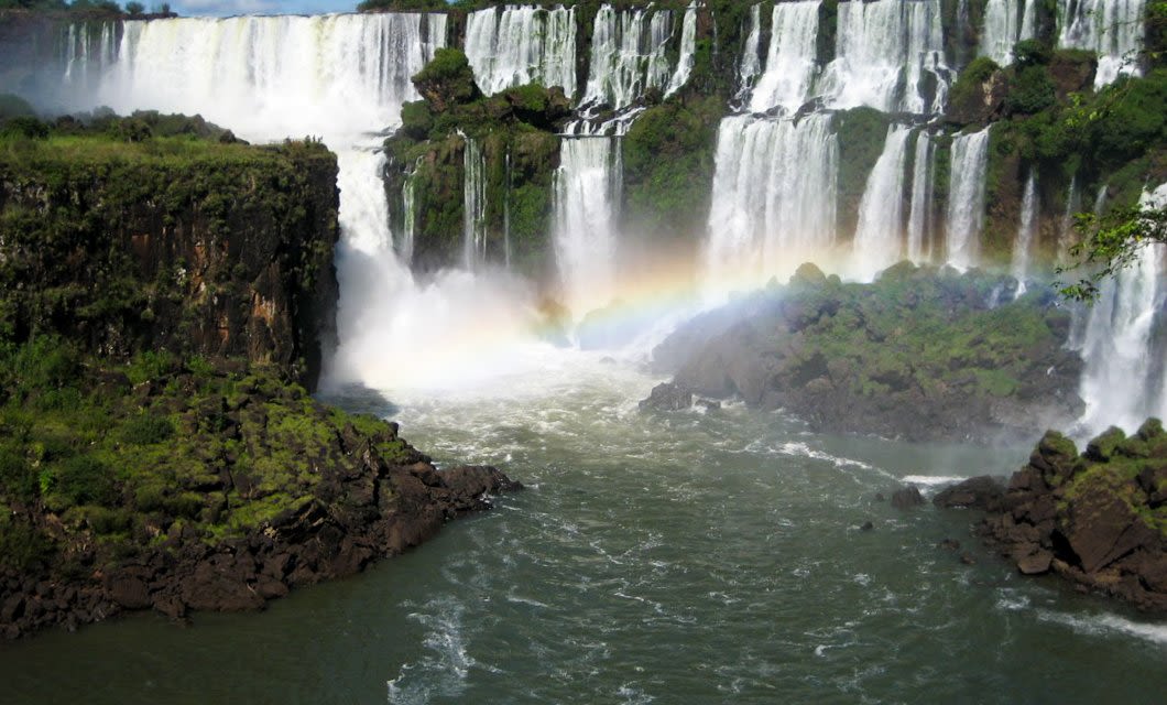 A wide river flows beneath tall, cascading waterfalls surrounded by lush greenery. Mist rises from the falls, and a faint rainbow arches across the scene near the water’s edge, evoking the spectacular beauty of Iguazu Falls.