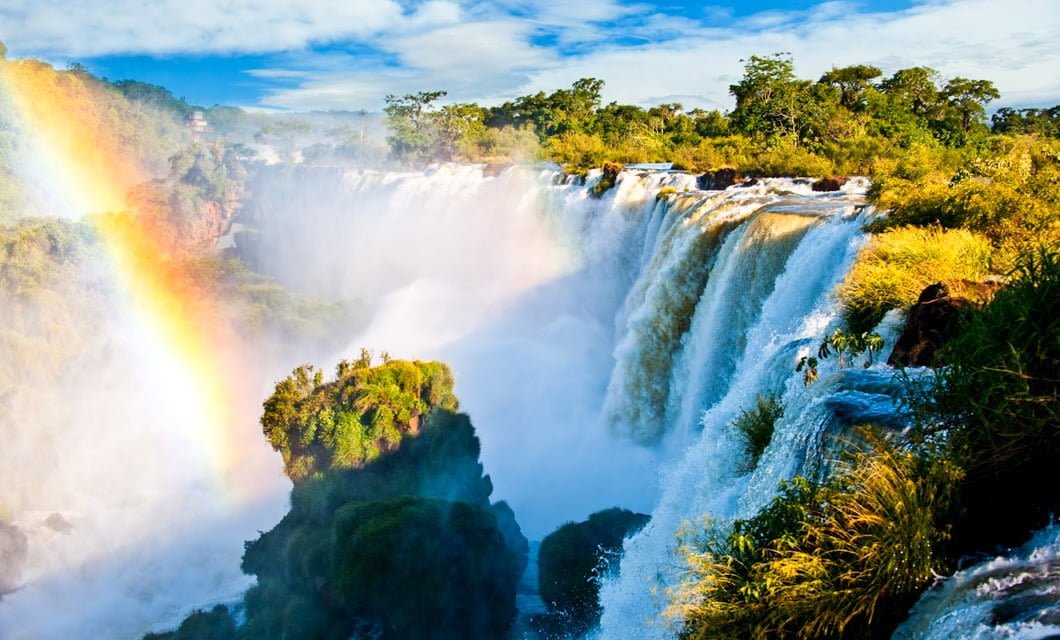 A powerful waterfall cascades over lush, green rocks with mist rising into the air. A bright rainbow arcs through the spray as dense forest surrounds the scene, reminiscent of landscapes once traversed by Inca explorers under a blue sky with scattered clouds.