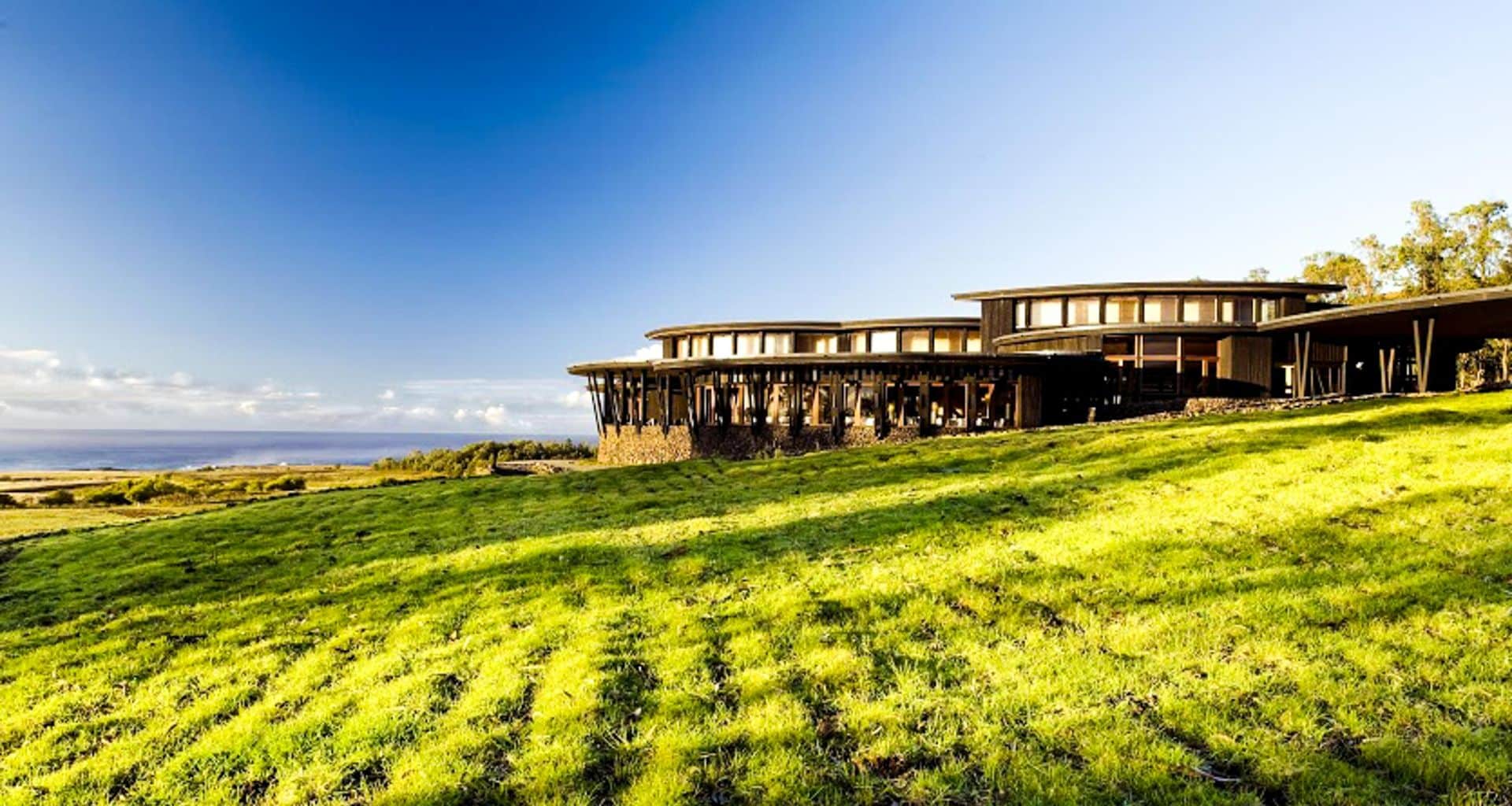 A modern, circular building with large windows sits on a grassy hillside under a clear blue sky at Explora Rapa Nui, with distant ocean and land visible in the background.