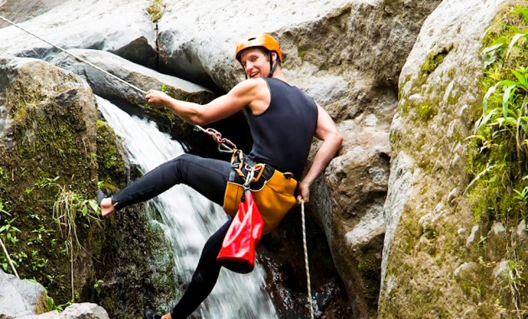 A person wearing a helmet and harness is rappelling down a rocky waterfall in the Atlantic Rainforest, surrounded by mossy rocks and greenery, using a rope for support on a thrilling Brazil Adventure Tour.