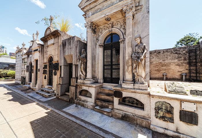 A row of ornate stone mausoleums with arched doorways and sculpted statues stands in a sunlit cemetery, with plaques and engraved details visible on the tombs, set not far from tranquil lakes that add serenity to the solemn scene.
