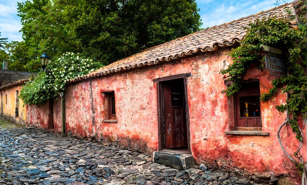 A rustic, weathered building with faded red walls and a tiled roof stands beside a cobblestone street in Colonia, surrounded by greenery and flowering plants under a partly cloudy sky.