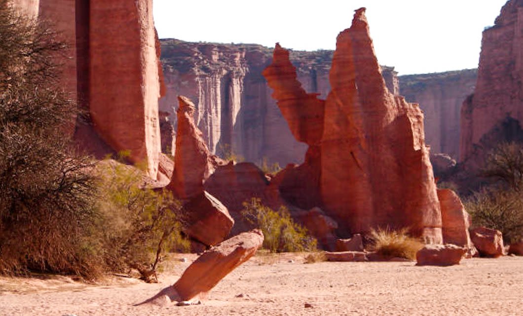Towering reddish rock formations rise from a dry, sandy landscape with sparse shrubs, standing as silent markers along the Boundaries of Time, set against imposing cliffs under a clear sky.