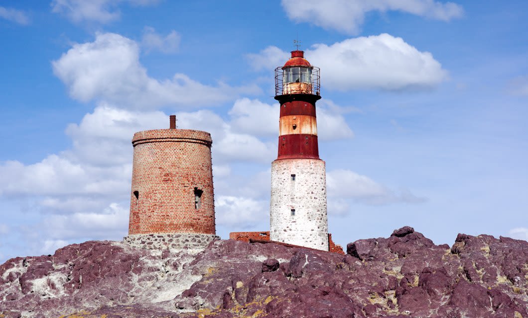 Two old brick lighthouses stand on rocky terrain in Puerto Deseado, Patagonia, under a partly cloudy sky—a perfect spot for bird watching by the rugged coast.