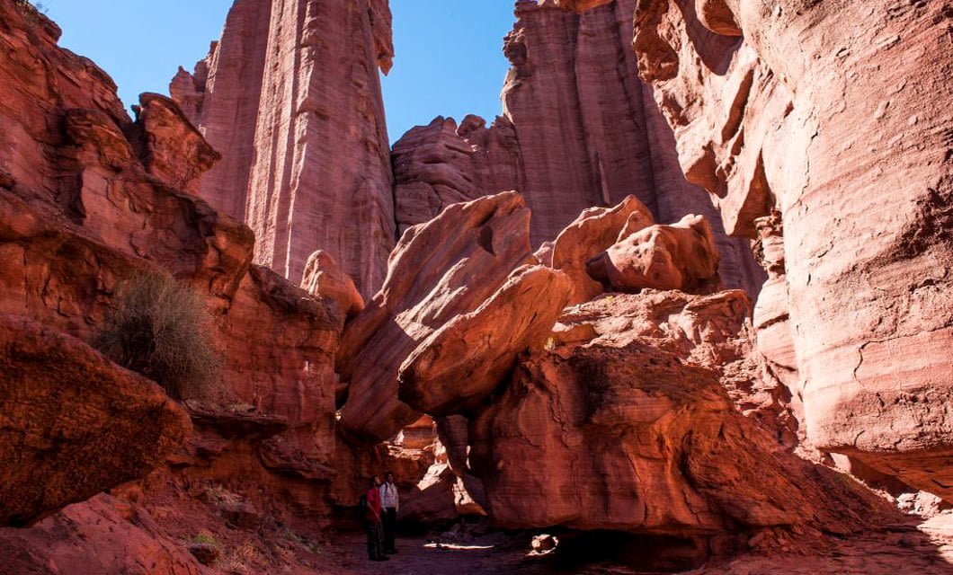 A person stands at the base of towering red rock formations and cliffs under a clear blue sky in a dramatic desert canyon landscape, reminiscent of scenery found in Triassic Parks.