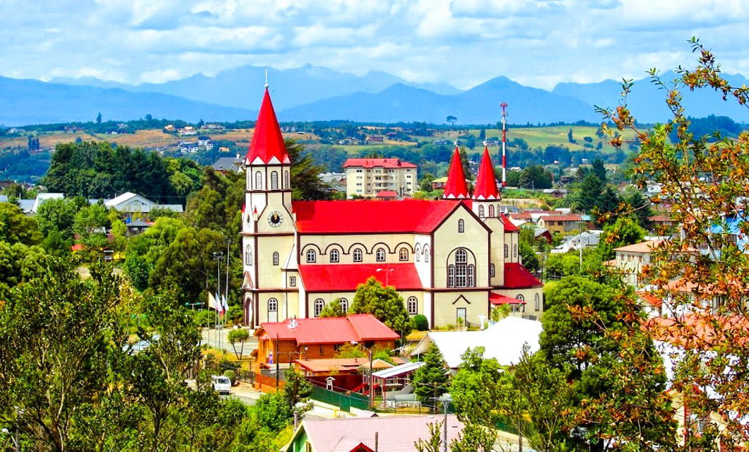 A large church with red roofs and spires stands among trees and houses in Patagonia, with mountains and a partly cloudy sky in the background. The Chilean town is surrounded by lush greenery.