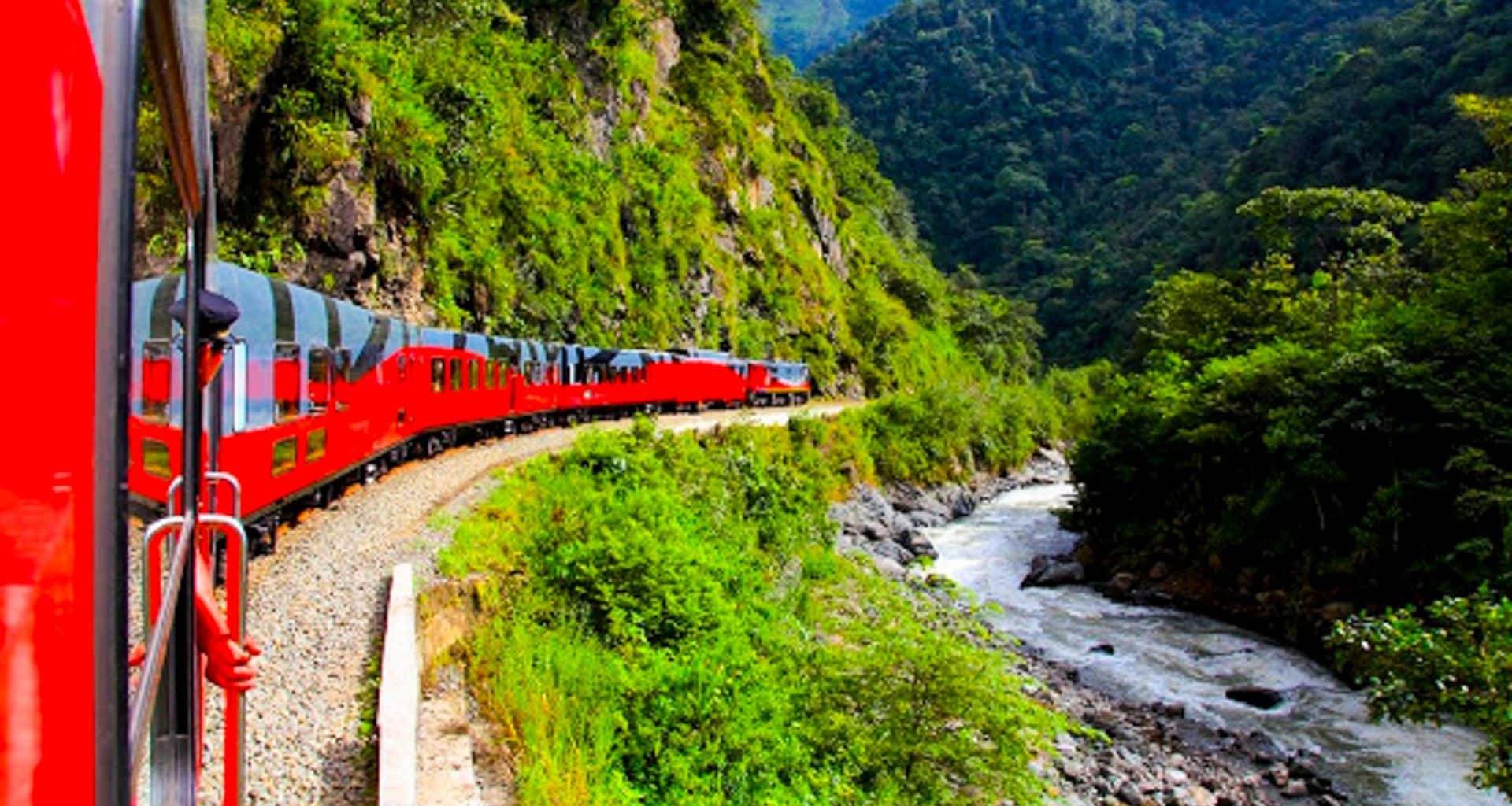 A bright red train embarks on a scenic train journey from Guayaquil to Quito, curving through lush green mountains with a fast-flowing river beside the tracks, under a vibrant blue sky.