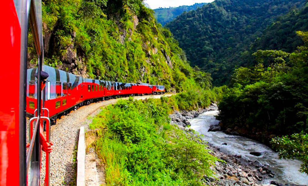 A bright red train winds along a curved track through lush green mountains beside a rushing river on the scenic Guayaquil to Quito train journey, with a passenger’s hand visible out the window.