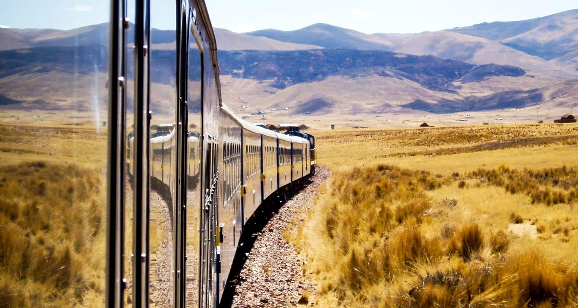 A long train, part of a scenic train tour, curves through a dry, grassy landscape in Bolivia with mountains in the background under a clear blue sky.